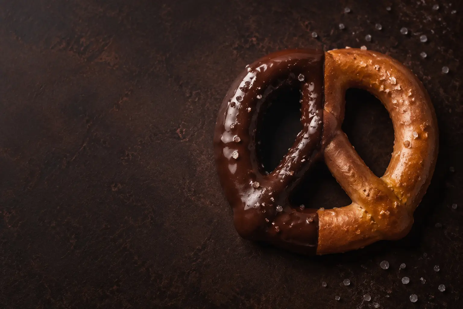 A real handmade pretzel on dark cocoa stone with one side dipped in glossy chocolate along a clean vertical boundary
